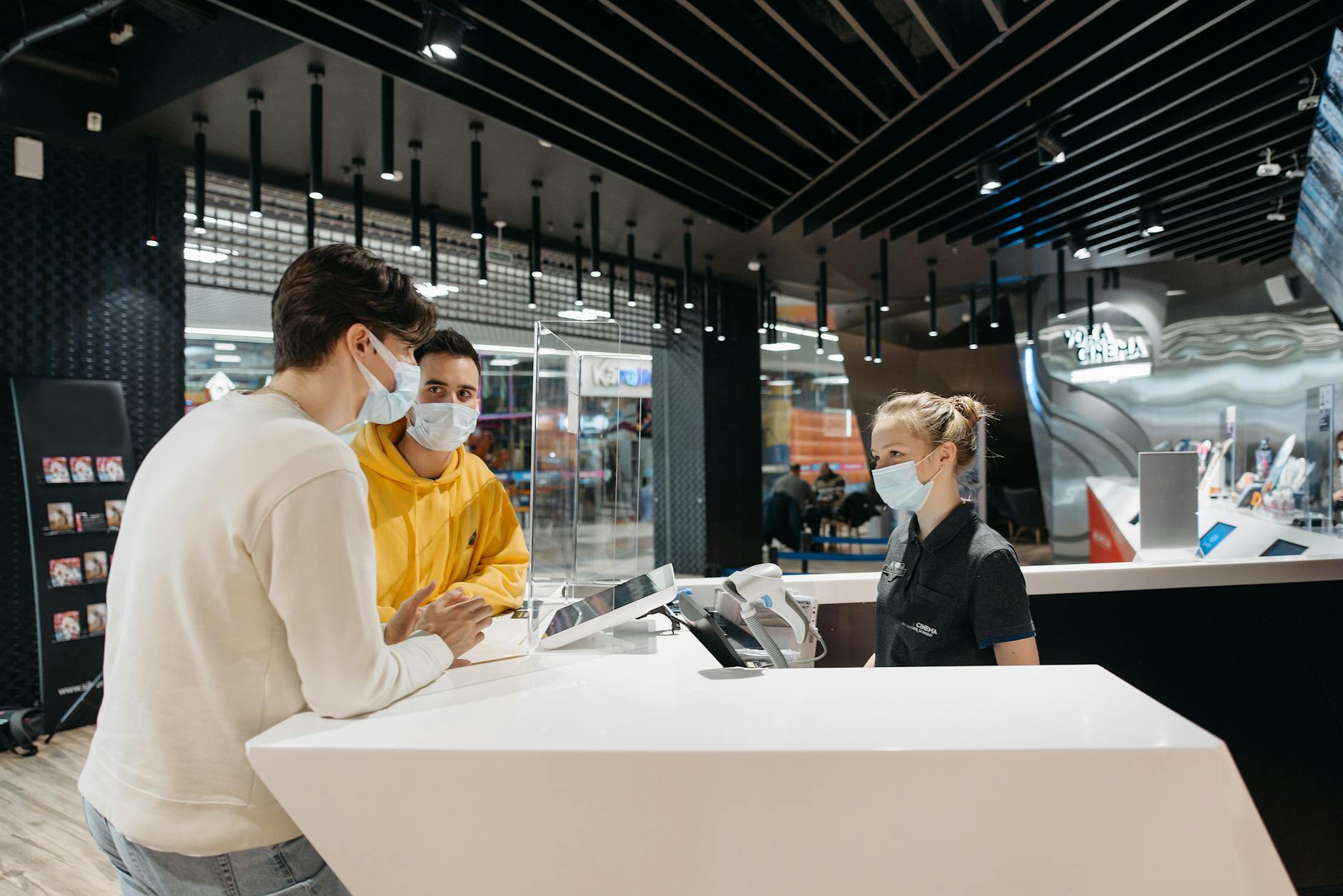 Two masked customers at a modern store counter interacting with a masked female staff member.